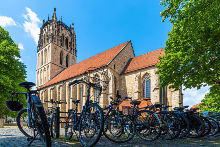 Vor einer historischen Steinkirche mit hohem Turm im Kreuzviertel von Münster stehen Fahrräder – der perfekte Ausgangspunkt für ein Outdoor-Escape-Game an einem sonnigen Tag.