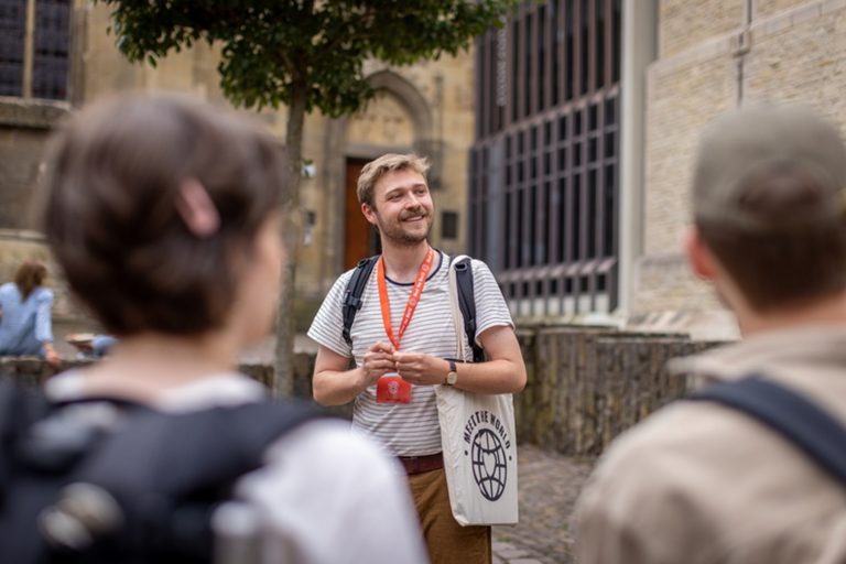 Ein lächelnder Reiseleiter mit einem roten Schlüsselband spricht vor einem historischen Gebäude zu einer Gruppe.