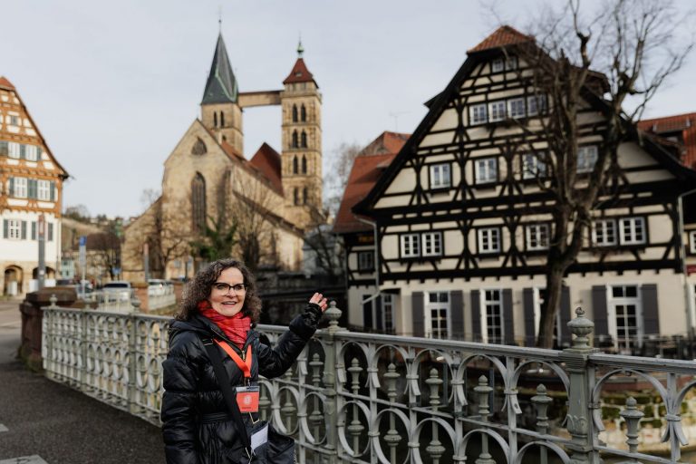 Frau in schwarzer Jacke zeigt auf historische Fachwerkhäuser und eine Kirche in Esslingen Mitte-West, perfekt für eine Kulinarische Stadtführung im Herzen von Esslingen.