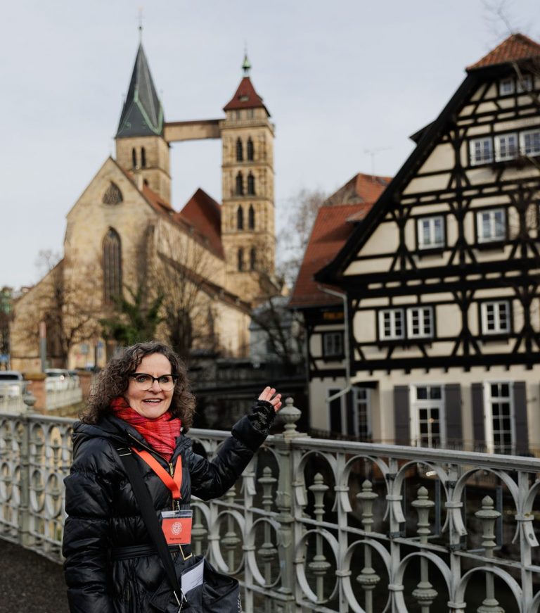 Eine Frau in einem schwarzen Mantel zeigt auf historische Gebäude und eine Kirche mit Türmen im Hintergrund und unterstreicht damit den Charme von Esslingen Mitte-West – einem Viertel, das oft im Rahmen einer kulinarischen Stadtführung durch Esslingen erkundet wird.