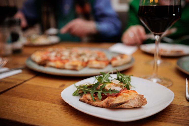 Ein Stück Pizza mit Rucola auf einem Teller, mit Wein und einer ganzen Pizza im Hintergrund - perfekt zum Genießen bei einer kulinarischen Stadtführung in Esslingen Mitte-West.