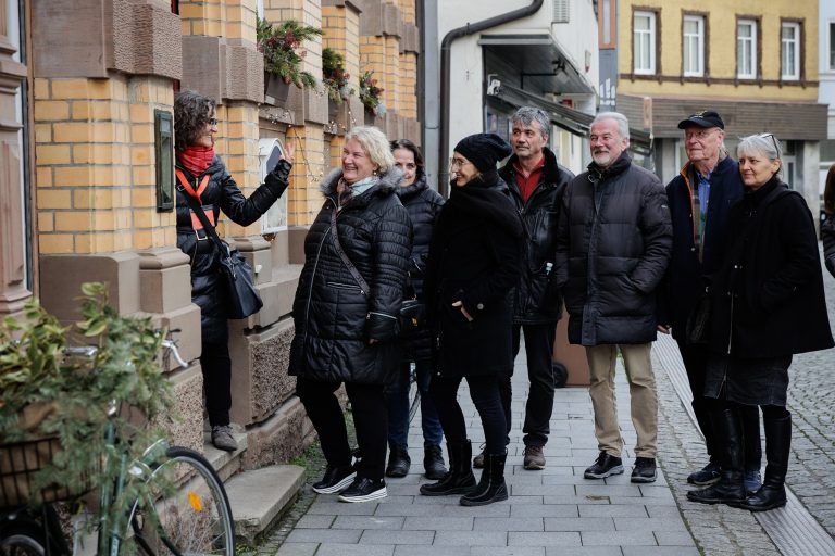 Eine Gruppe von Menschen in Wintermänteln lächelt einer Frau zu, die sie während der Kulinarischen Stadtführung in Esslingen Mitte-West auf einem Bürgersteig fotografiert.
