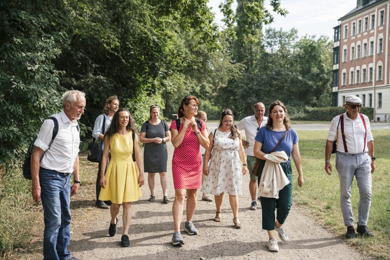 Eine Gruppe von Menschen genießt eine Stadtführung und schlendert an einem sonnigen Tag einen von Bäumen gesäumten Weg in der Nähe eines Gebäudes im Südviertel von Münster entlang.