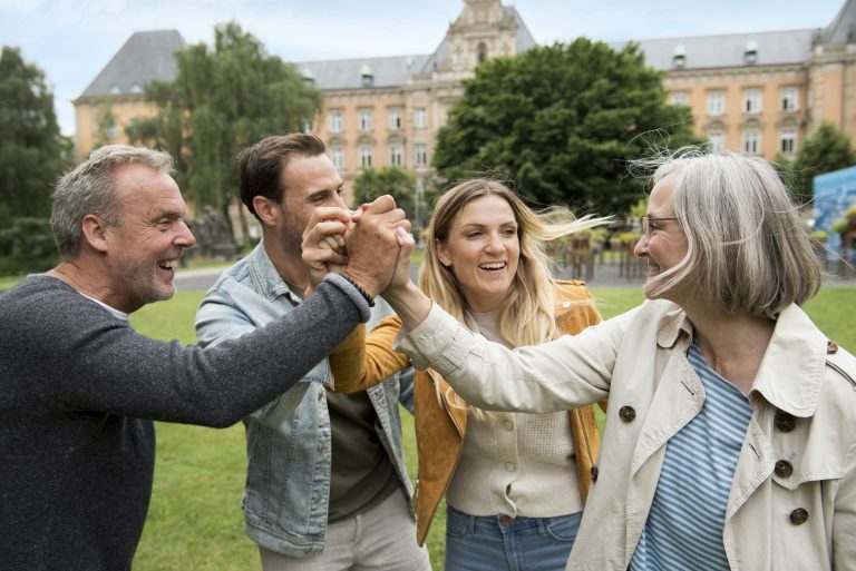 Vier Menschen lächeln und geben sich im Freien ein High Five, mit einem historischen Gebäude und Bäumen im Hintergrund, und genießen eine der vielen Aktivitäten, die Hamburg zu bieten hat.