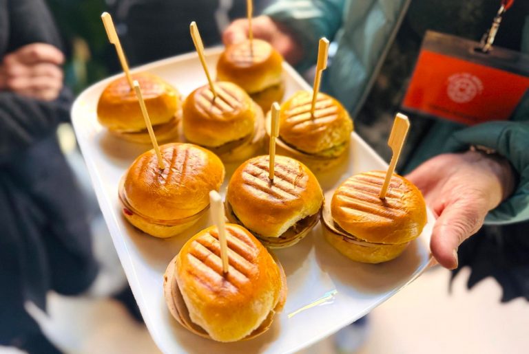 Eine Person hält ein Tablett mit kleinen gegrillten Sliders, die jeweils mit einem Holzspieß befestigt sind - perfekte Happen für eine kulinarische Stadtführung durch den Prenzlauer Berg.