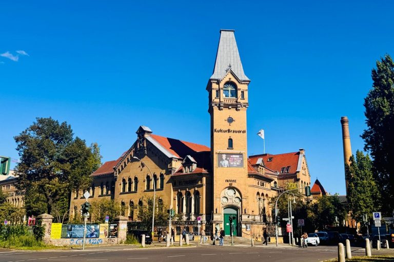 Historisches Backsteingebäude mit hohem Uhrenturm, rotem Dach und "KulturBrauerei"-Schild unter strahlend blauem Himmel - ein Wahrzeichen des Prenzlauer Bergs, das häufig bei kulinarischen Stadtführungen in Berlin zu sehen ist.