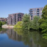 Moderne Backstein-Wohnhäuser an einem ruhigen Fluss im Hamburger Stadtteil Barmbek, umgeben von üppigen grünen Bäumen unter einem klaren blauen Himmel.