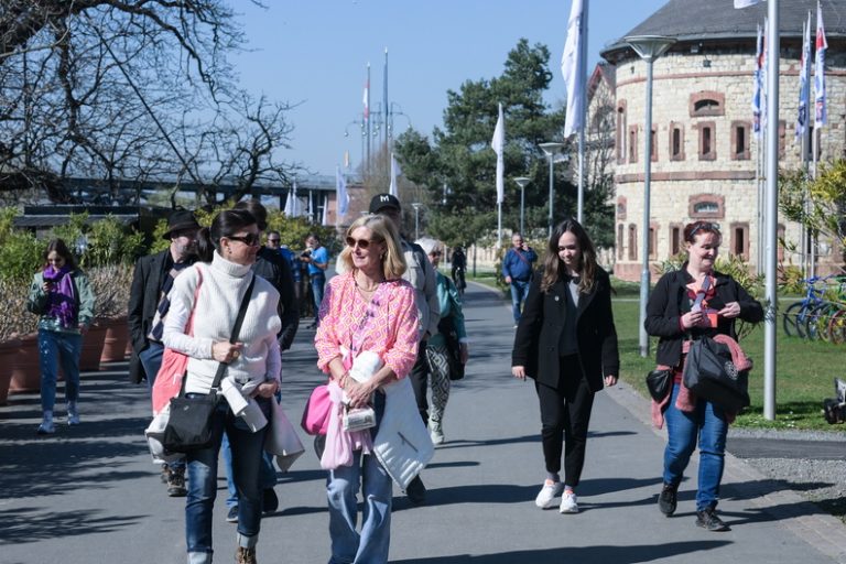 Eine Gruppe von Menschen geht auf einem sonnigen Weg in der Nähe eines Backsteingebäudes mit Bäumen und Fahnen im Hintergrund und genießt eine Aktiv-Tour Mainz im Rahmen einer herrlichen Kulinarischen Tour.