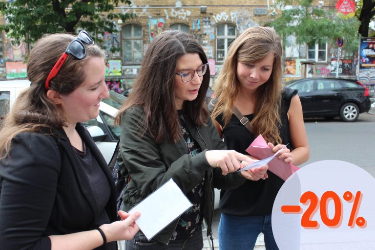 Drei Frauen diskutieren im Freien über aktuelle Artikel und erkunden die neuesten Frühlingshighlights. Eine „-20%“-Grafik in der unteren rechten Ecke weist auf unwiderstehliche Angebote hin.