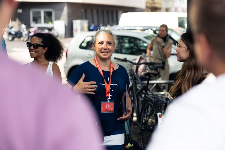 Eine Frau mit einem roten Schlüsselband spricht zu einer Gruppe im Freien während einer kulinarischen Stadtführung in Köln, in der Nähe geparkter Fahrräder und Autos an einem sonnigen Tag.