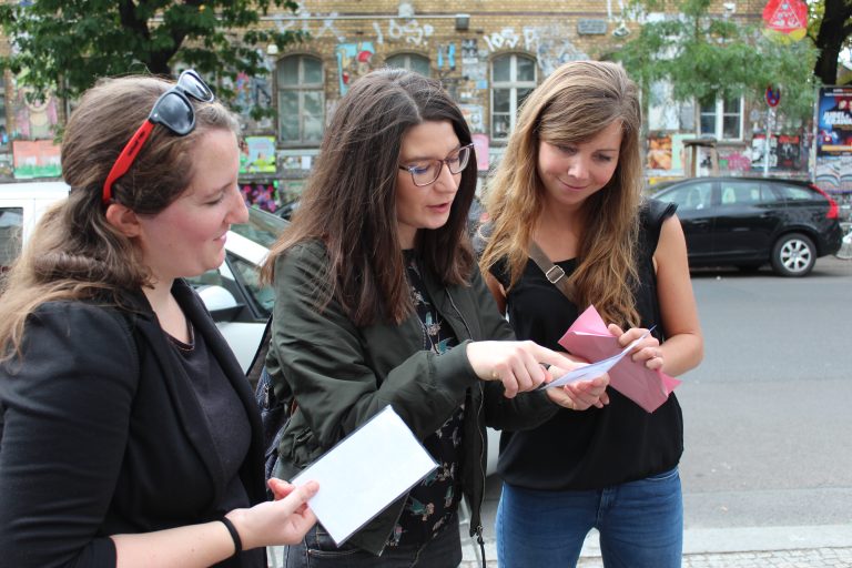 Drei Frauen stehen auf einer belebten Straße und diskutieren angeregt über Frühlingshighlights, während sie gemeinsam auf ein Blatt Papier starren.