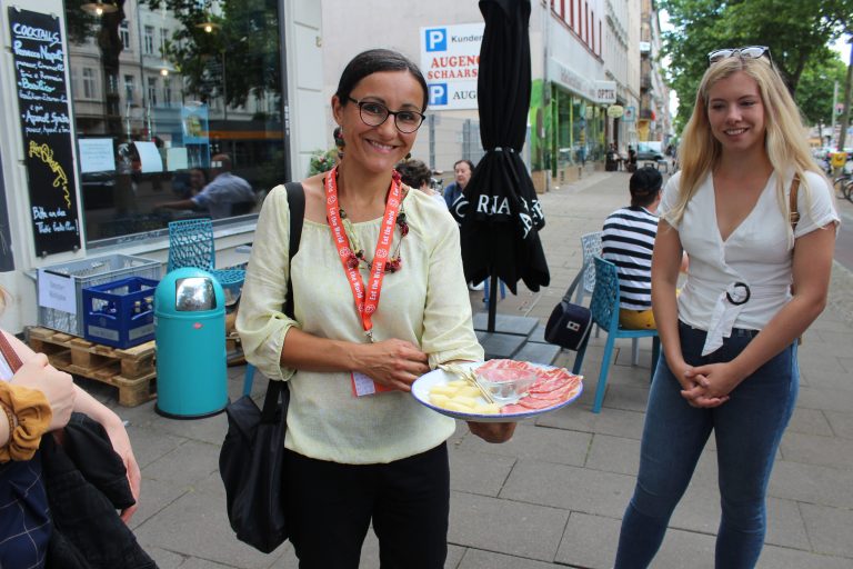 Vor einem charmanten Café präsentiert eine Frau freudig einen Teller mit Aufschnitt und Käse und teilt den Genuss der Frühlingshighlights mit einer anderen lächelnden Frau an ihrer Seite.