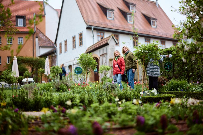 Drei Frauen stehen lächelnd in einem üppigen Garten in Weimar, mit dem historischen Gebäude des Quartier der Moderne im Hintergrund.