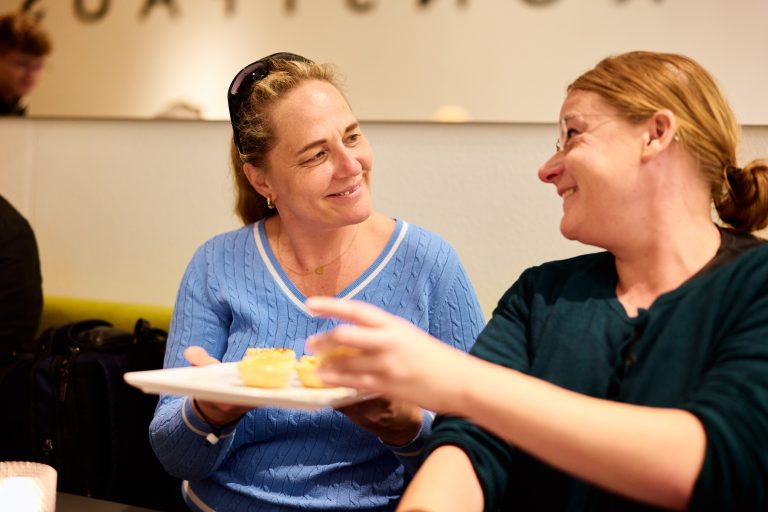 Zwei Frauen, die lächelnd in einem Innenraum in Weimar zusammen essen, eine hält einen Teller mit Gebäck in der Hand - sie fangen die Freude an einer kulinarischen Stadtführung durch die Aromen des Quartiers der Moderne ein.