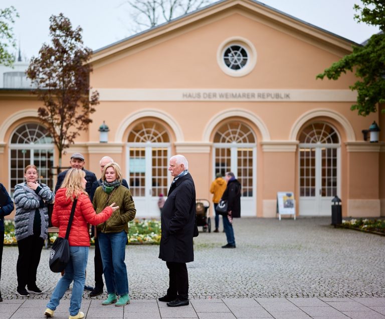 Menschen unterhalten sich vor dem Haus der Weimarer Republik, einem beigen Gebäude mit Rundbogenfenstern und Blumen im Außenbereich im Weimarer Quartier der Moderne.