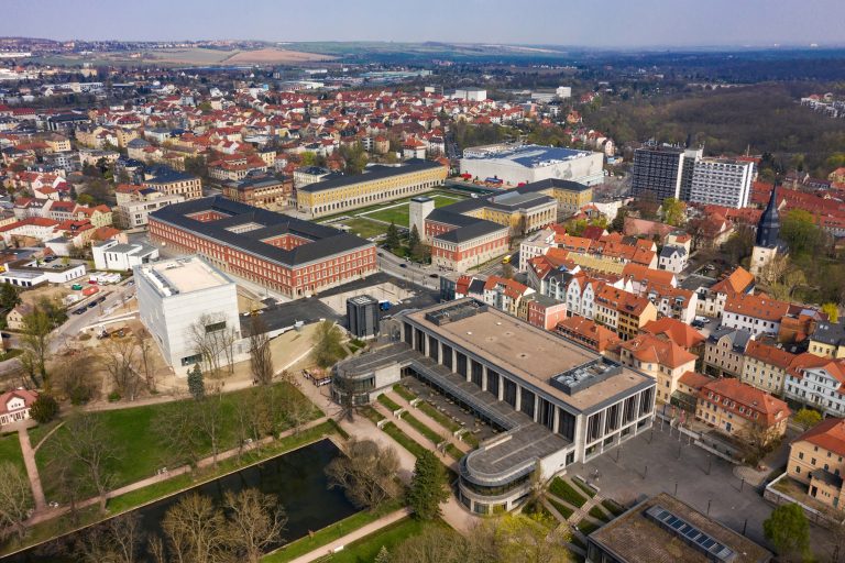 Luftaufnahme von Weimar mit modernen und historischen Gebäuden, einschließlich des Quartiers der Moderne, üppigen Parks und entfernten Hügeln unter klarem Himmel.