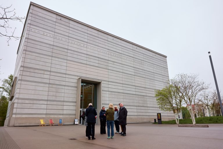 Eine Gruppe von Menschen steht vor dem Quartier der Moderne, einem großen, modernen grauen Betongebäude in Weimar, bereit, an einem bewölkten Tag ihre kulinarische Stadtführung zu beginnen.