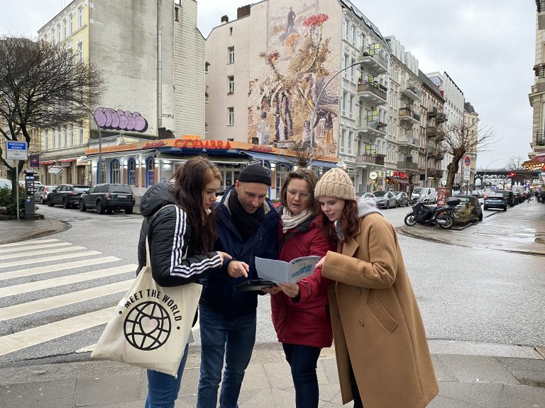 Vier Personen stehen an einer Berliner Straßenecke und schauen gemeinsam auf eine Karte, während sie an einem bewölkten Tag ihren nächsten Schritt bei einer Schnitzeljagd planen.