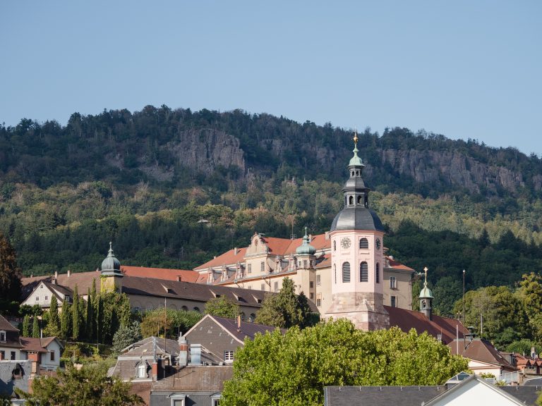 Eine rosafarbene Kirche mit einem hohen Turm steht zwischen Bäumen im Oostal, im Hintergrund ein bewaldeter Hügel bei klarem Himmel - die perfekte Kulisse für eine Kulinarische Aktiv-Tour bei Baden-Baden.