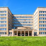Ein großes hellbraunes Universitätsgebäude mit vielen Fenstern steht auf einer Wiese unter strahlend blauem Himmel und erinnert an den architektonischen Charme des Frankfurter Westends.