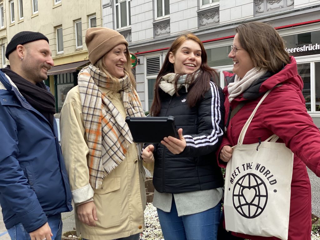 Vier Personen in Winterkleidung lächeln und schauen auf eine Tafel im Freien auf einer Stadtstraße und genießen eine gemeinsame Stadtführung zu Fuß.