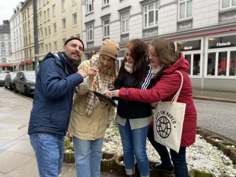 Vier Personen in Winterkleidung schauen gemeinsam auf ein Tablet auf einem städtischen Gehweg in der Nähe eines Blumenbeets und suchen nach spannenden Aktivitäten, die Bremen zu bieten hat.