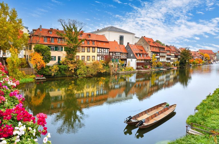 Bunte Häuser und Blumen säumen einen ruhigen Fluss in der Bamberger Gärtnerstadt, wo zwei Holzboote unter einem hellen, teilweise bewölkten Himmel ruhen - perfekt für den Beginn einer kulinarischen Stadtführung.
