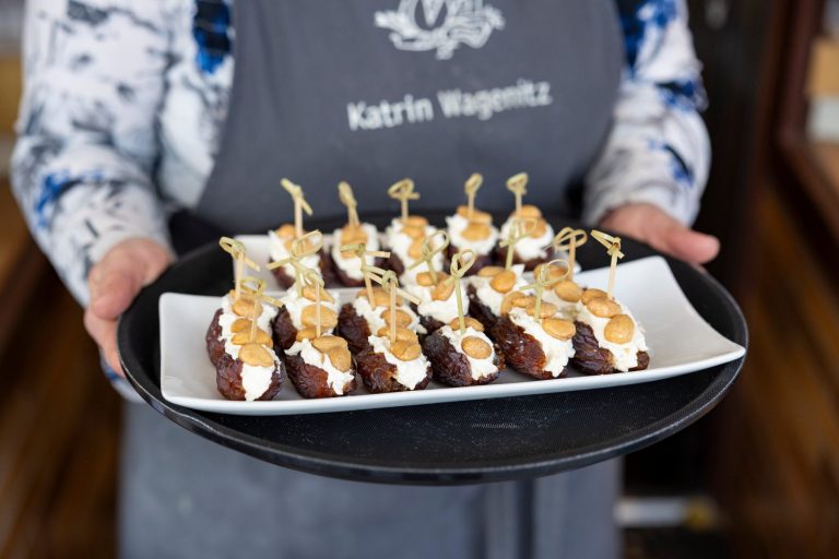 Während einer kulinarischen Stadtführung in der Bremer Neustadt hält eine Person einen Teller mit Datteln, die mit Sahne gefüllt und mit Nüssen belegt sind, jeweils mit einem kleinen Spieß.