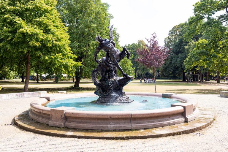 Ein Bronzebrunnen in einem runden Becken, umgeben von Bäumen, im Park der Bremer Neustadt an einem sonnigen Tag, ideal zum Entspannen nach einer kulinarischen Stadtführung.