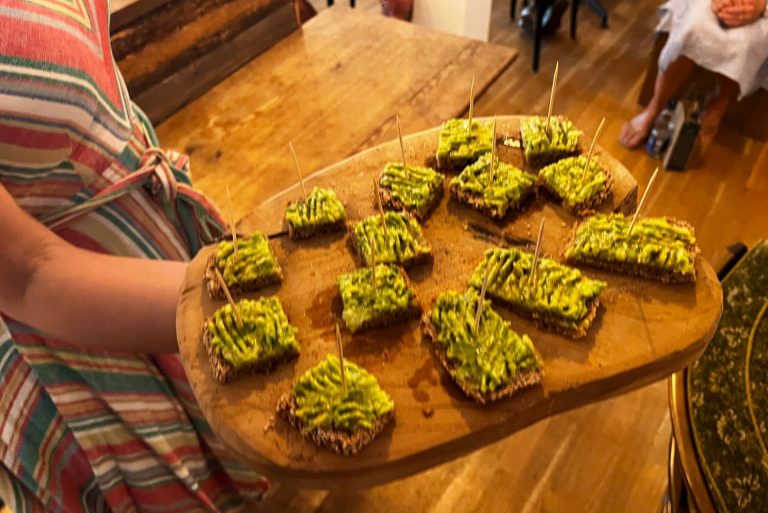 Eine Person, die in einem Innenraum ein Holzbrett mit mehreren Avocado-Toasts hält, die jeweils mit einem Zahnstocher gespickt sind – perfekt für eine kulinarische Stadtführung durch Tübingens charmante Altstadt.
