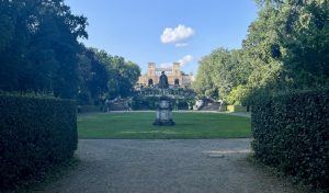 Eine Statue steht vor einem kunstvollen Gebäude, umgeben von Bäumen und Hecken, unter einem blauen Wolkenhimmel, der an den Charme der schönsten Schlösser und malerischen Parks erinnert, für die Potsdam bekannt ist.