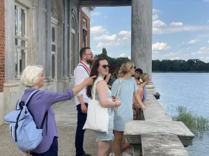 Eine Gruppe von Menschen steht an einem Gebäude am Seeufer und blickt bei teilweise bewölktem Himmel auf das Wasser. Sie genießen einen der schönen Parks, für die Potsdam bekannt ist.