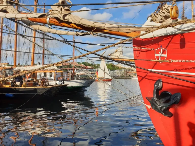 Nahaufnahme eines roten Segelboots mit Bug und Anker in der Flensburger Neustadt, wo weitere Segelboote und Wasserspiegelungen den Charme einer kulinarischen Stadtführung in diesem lebendigen Hafenviertel ausmachen.