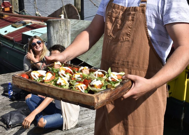 Eine Person in Schürze hält ein Tablett mit Häppchen in der Hand; zwei Personen sitzen am Wasser in der Flensburger Neustadt, eine zeigt den Daumen nach oben während einer kulinarischen Stadtführung.