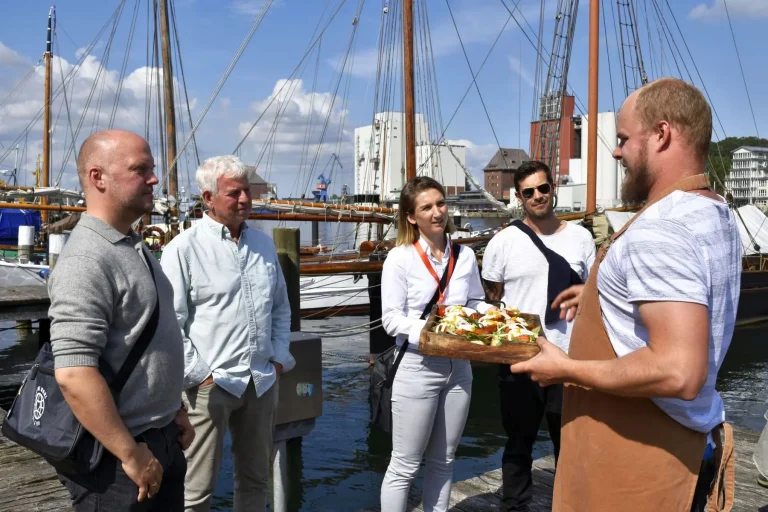 Ein Koch präsentiert vier Personen auf einem Steg in der Flensburger Neustadt ein Tablett mit Speisen. Im Hintergrund sind Segelboote zu sehen, die den Charme einer kulinarischen Stadtführung am Hafen versprühen.