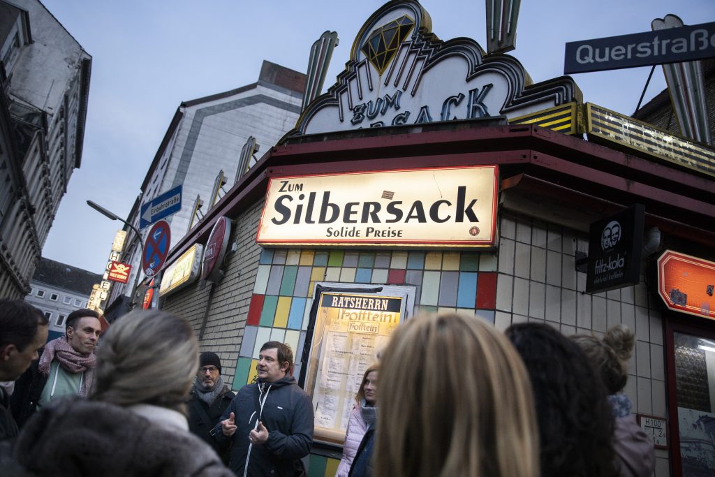Eine Gruppe von Menschen versammelt sich in der Abenddämmerung vor der Bar Zum Silbersack mit einem hell erleuchteten Schild in einer Stadtstraße - eine der klassischen Date Ideen in Hamburg.