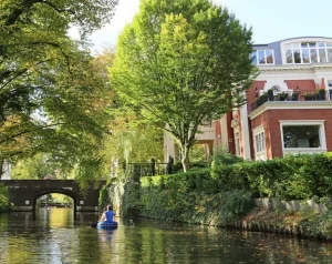 Eine Person fährt mit dem Kajak auf einem Kanal neben einem großen roten Backsteinhaus und üppig grünen Bäumen und genießt eine der einzigartigen Date-Ideen, die Hamburg zu bieten hat.