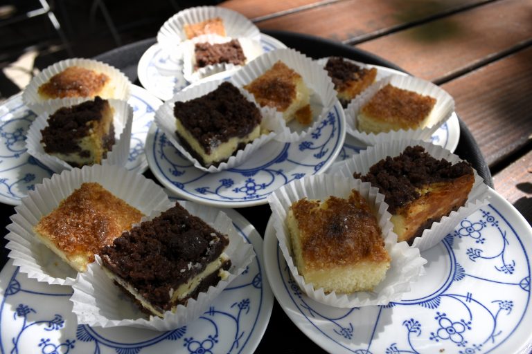 Neun Teller mit verschiedenen Kuchenscheiben in Papiereinlagen auf einem Tablett, Sonnenlicht wirft Schatten auf den Tisch - perfekt für einen Entdecker-Genießer-Tag in Potsdam.