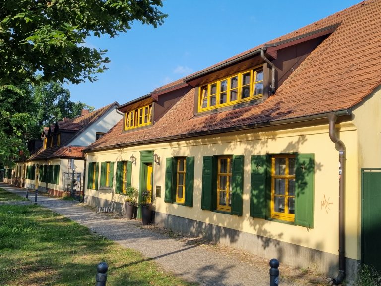Sonnige Straße in Potsdam mit einem gelben Haus, grünen Fensterläden, rotem Dach und schattenspendenden Bäumen - perfekt für Entdecker und Genießer, um einen entspannten Tag zu genießen.
