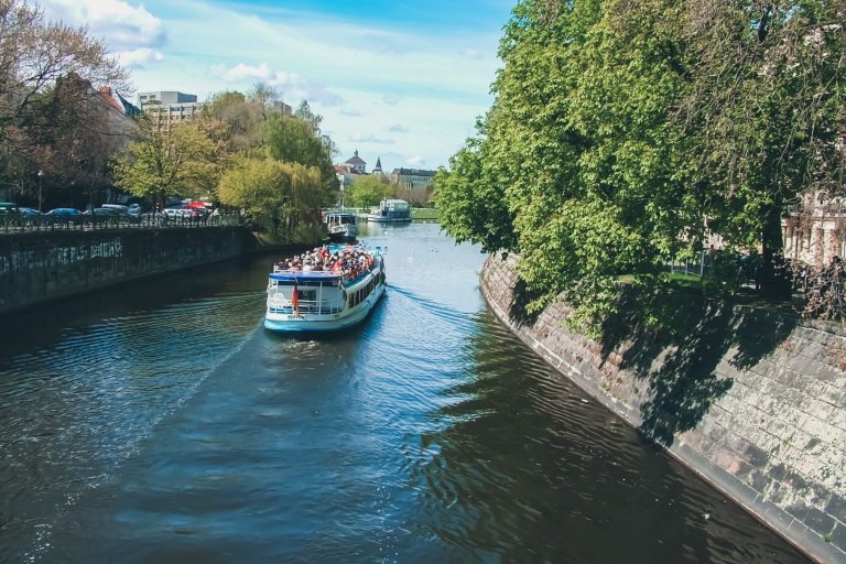 Ein Ausflugsboot mit Entdecker und Genießer fährt an einem sonnigen Tag den baumbestandenen Kanal in Potsdam entlang.