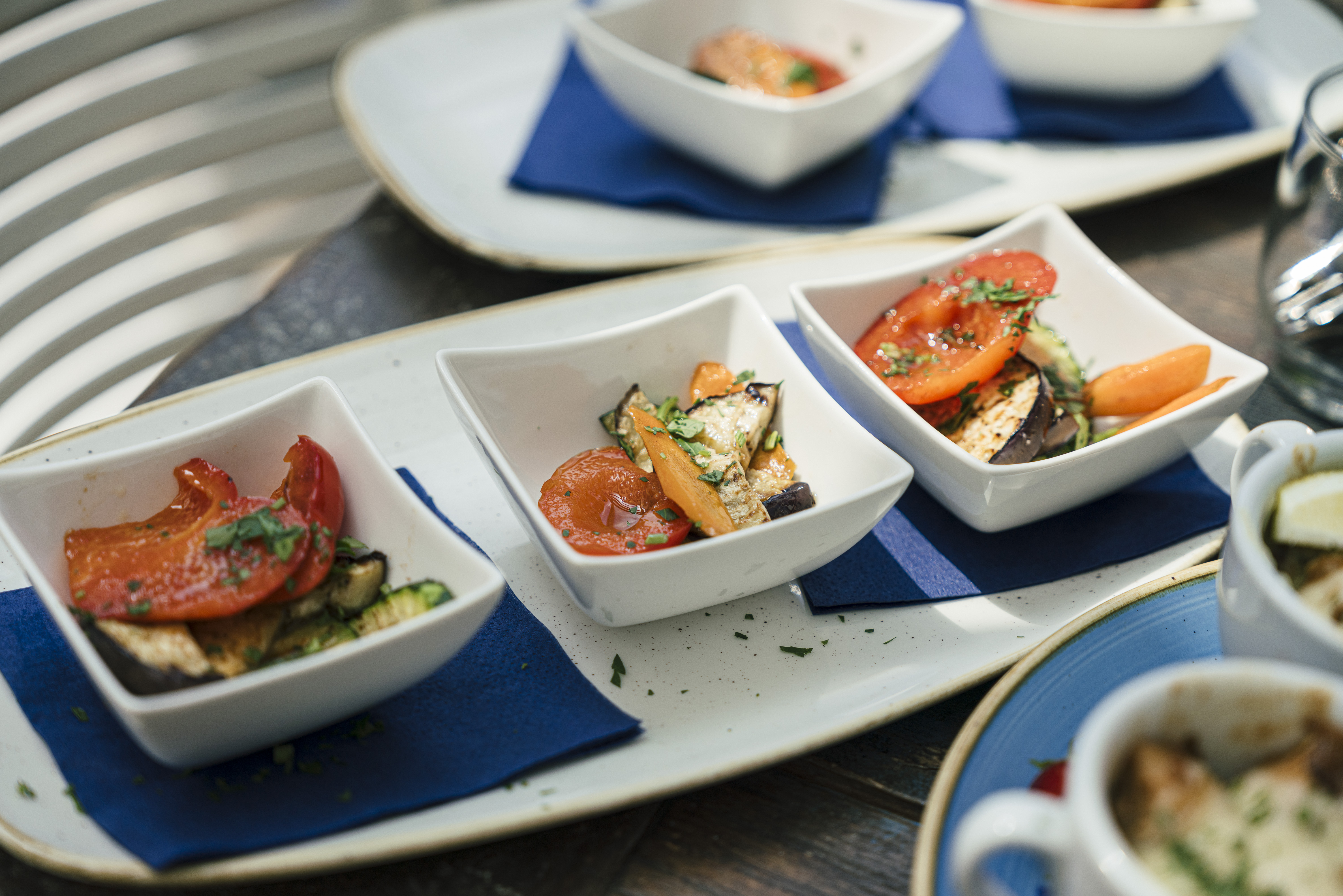 Drei kleine Schälchen mit gegrilltem Gemüse, hauptsächlich Tomate und Zucchini, serviert auf blauen Servietten auf einem weißen Tablett - perfekt für schöne Dates in Leipzig oder als leckerer Snack während Ihrer Leipziger Unternehmungen.
