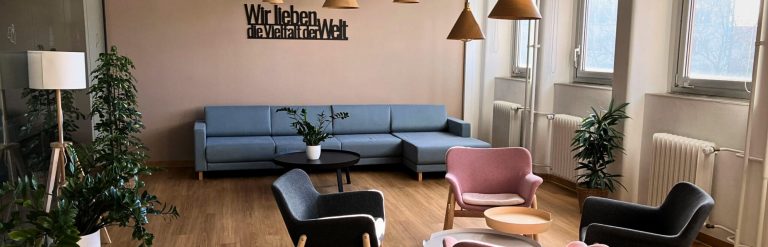 Modernes Wohnzimmer mit blauem Sofa, rosafarbenen und grauen Sesseln, Pflanzen, Holzfußboden und deutschsprachiger Wandmalerei, ideal für Telearbeit.
