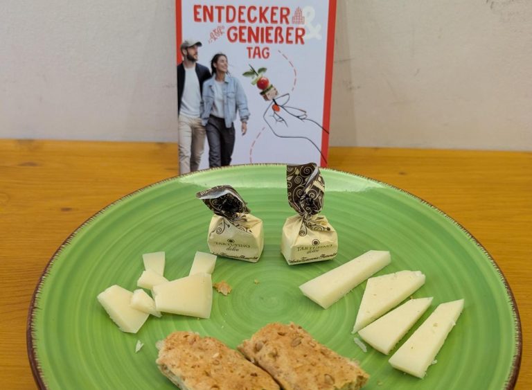 Ein grüner Teller mit Käsescheiben, zwei eingewickelten Bonbons und Biscotti steht bereit für einen „Entdecker-Genießer-Tag“ in Heidelberg; im Hintergrund ist ein deutscher Flyer zu sehen.