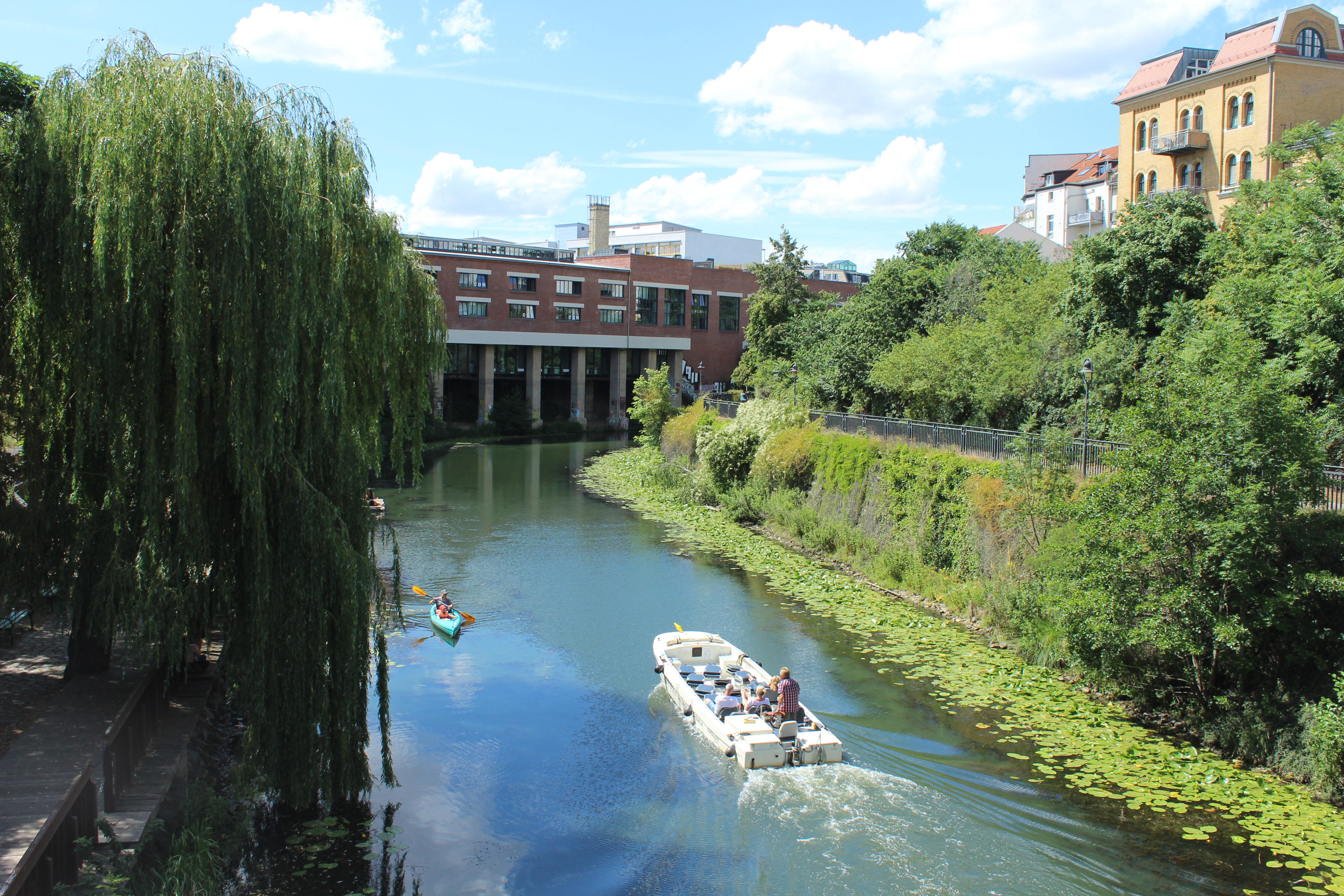 Ein weißes Boot und ein Kajak fahren an einem sonnigen Tag einen von Bäumen und Gebäuden gesäumten Kanal hinunter - einer von vielen schönen Terminen, die Leipzig für diejenigen zu bieten hat, die einzigartige Stadterlebnisse suchen.