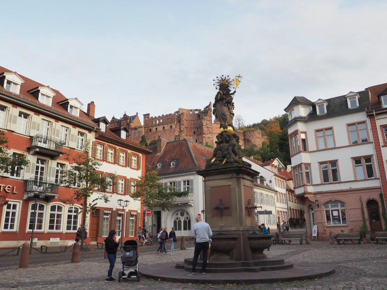 Entdecker und Genießer versammeln sich um eine Statue auf einem europäischen Platz, während im Hintergrund die Silhouette des Heidelberger Schlosses den Tag krönt.