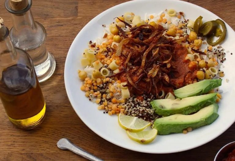 Ein Teller Koshari mit Reis, Linsen, Nudeln, knusprigen Zwiebeln, Avocado, Limette und Soßen auf einem Holztisch im pulsierenden Wrangelkiez - perfekt für eine kulinarische Stadtführung.