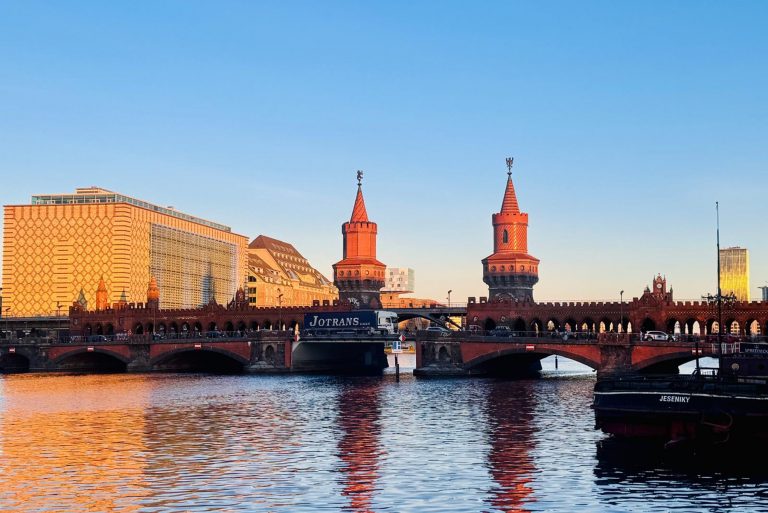 Eine rote Ziegelsteinbrücke mit zwei Türmen überspannt einen Fluss in Berlin, mit städtischen Gebäuden und blauem Himmel im Hintergrund, in der Nähe des pulsierenden Wrangelkiezes.