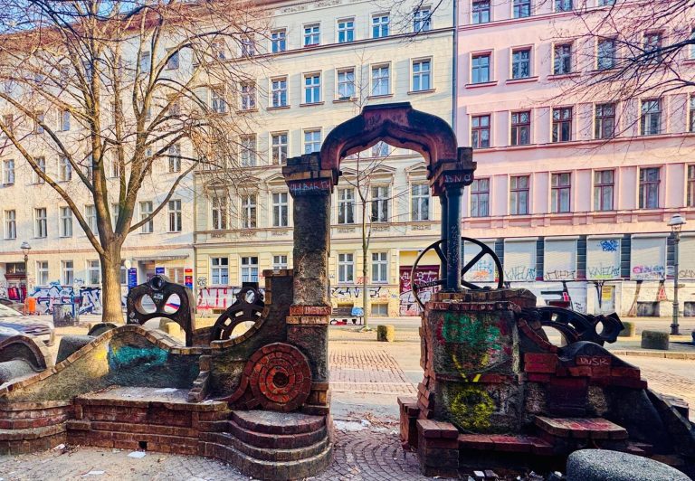 Bunte, verwitterte Steinbogen-Skulpturen auf einem Stadtplatz im Berliner Wrangelkiez, mit bemalten Wohnhäusern und Bäumen dahinter.