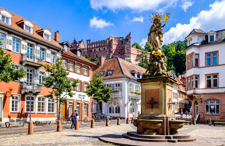 Bunte Gebäude und eine Statue auf einem kopfsteingepflasterten Platz laden Entdecker zum Entdecken ein, während im Hintergrund das Heidelberger Schloss auf dem Hügel thront.