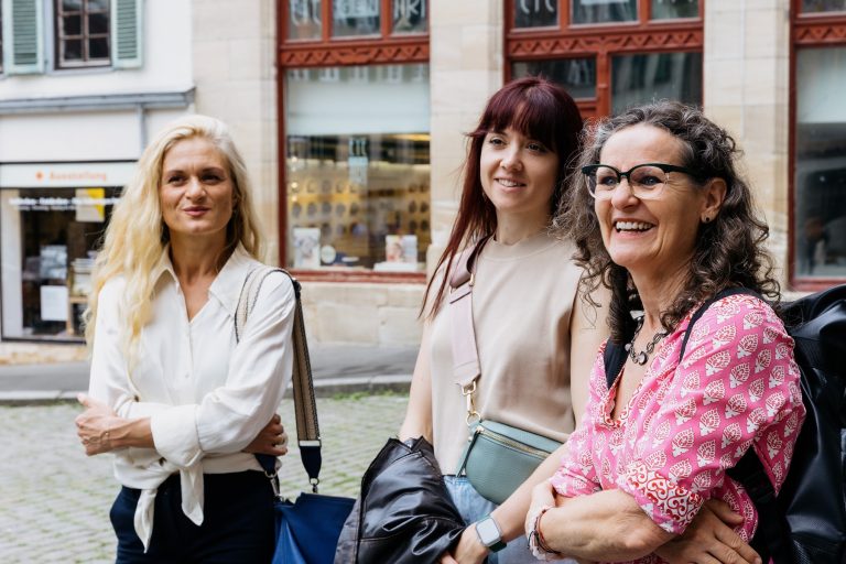 Drei Frauen stehen im Freien auf einer Heidelberger Straße, lächeln und unterhalten sich, genießen einen echter Entdecker-Genießer-Tag mit charmanten Geschäften im Hintergrund.
